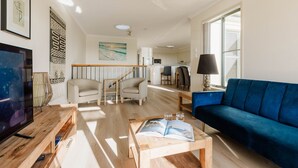 Ein Wohnzimmer mit einem blauen Sofa, einem Couchtisch mit Büchern, einem Fernseher und einer Treppe, die in eine Küche führt.