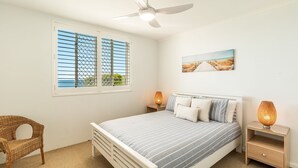 A bedroom with a bed, a ceiling fan, a window with blinds, a painting, and a nightstand with a lamp. - Craigmore 5 - Oceanfront Escape with Yamba's Best Beach Views - Holiday Yamba (Yamba)