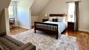 A bedroom with a sloped ceiling, a bed with a wooden headboard, a nightstand, a desk, and a chair. - Lamplight Cottage | paddle-boards, AC, best beach! (Sylvan Lake)