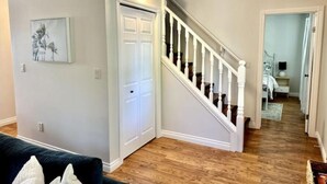 A spacious living room with hardwood floors, a staircase with white balusters, and a door leading to another room. - Lamplight Cottage | paddle-boards, AC, best beach! (Sylvan Lake)