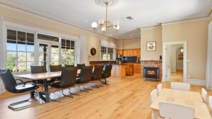 A spacious dining area with a long wooden table and modern chairs, a kitchen with wooden cabinets and a stone fireplace, and large windows offering a view of the outdoors. - Cornhill - Merrijig - Sleeps 12 (Merrijig)