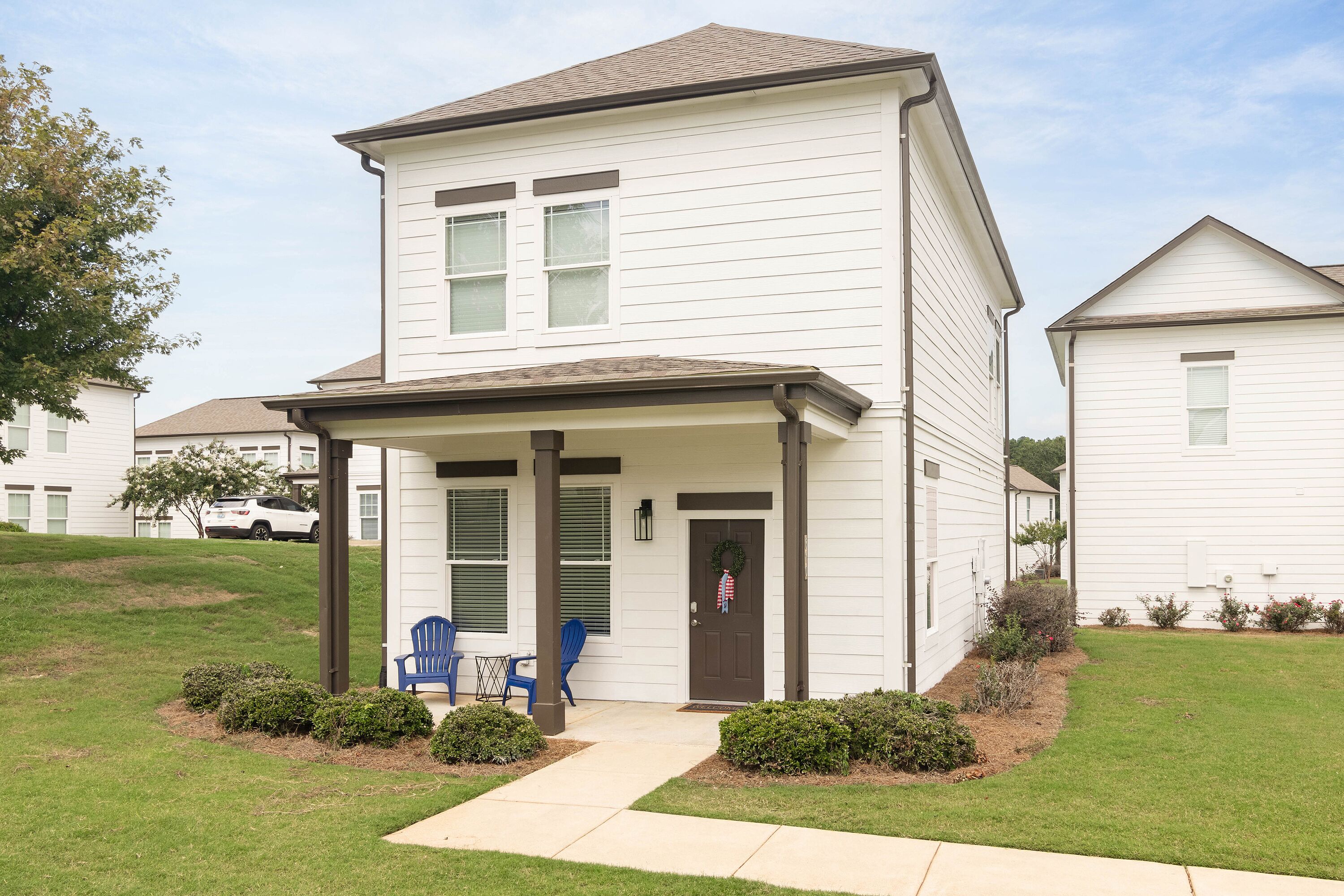 A two-story house with a covered porch, a front door with a wreath, and a small lawn area with bushes and a pathway.