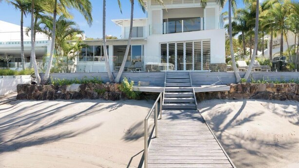 Un edificio moderno su due piani con un patio in legno che si apre su una spiaggia di sabbia, circondato da palme.