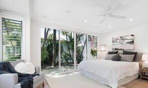 Une chambre à coucher comprenant un lit, une table de chevet, un ventilateur de plafond et une vue sur un jardin, visible à travers des portes vitrées.
