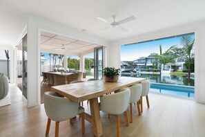 A modern dining area with a large wooden table and chairs, a ceiling fan, and a view of a swimming pool and buildings. - Lucy - Noosa - Noosaville (Noosaville)