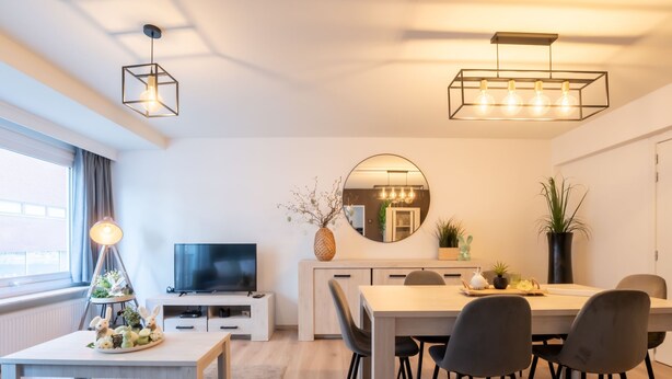A modern living room with a TV, dining table, and chairs. There is a large window with curtains, a round mirror, and a hanging light fixture. - Amuzee with parking & lateral sea view by Interhome (Blankenberge)
