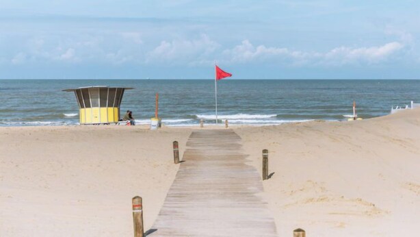 Een strand met een houten looppad dat leidt naar een rederspost en een rode vlag.