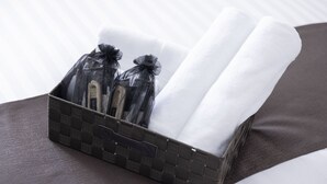 A hotel room with a neatly arranged set of toiletries in a woven basket, placed on a bed with a white pillow and a dark brown headboard. - Coruscant Hotel Nagasaki Station 3 (Nagasaki)