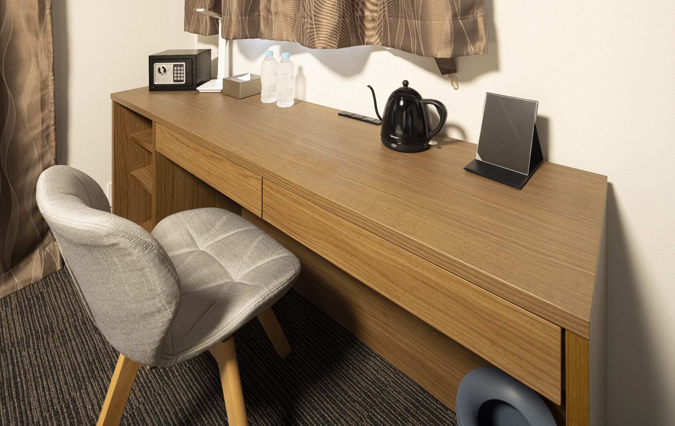 A wooden desk with a black kettle, a remote control, and a smartphone. There are two water bottles and a box on the desk. A grey chair is positioned in front of the desk.