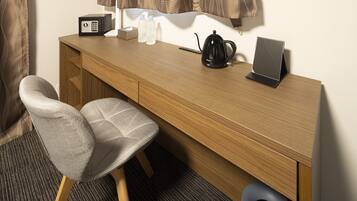A wooden desk with a black kettle, a remote control, and a smartphone. There are two water bottles and a box on the desk. A grey chair is positioned in front of the desk.