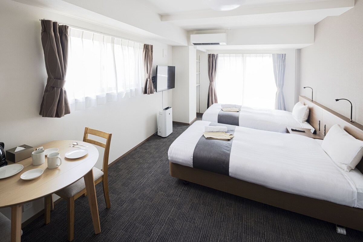 A hotel room with two beds, a TV, a desk with a chair, and a window with curtains.