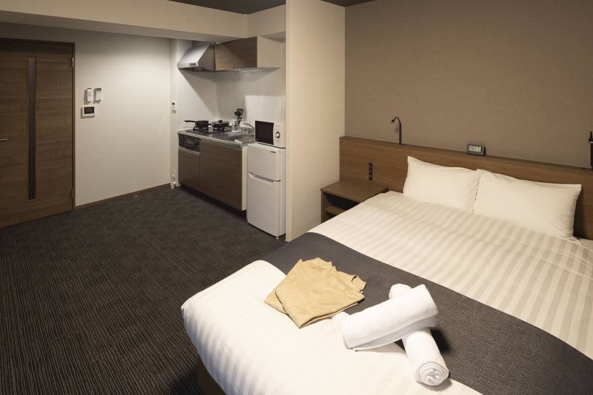Ein Hotelzimmer mit Bett, einer kleinen Küchenzeile, einem Holz-Kopfteil und einem Nachttisch mit Lampe.