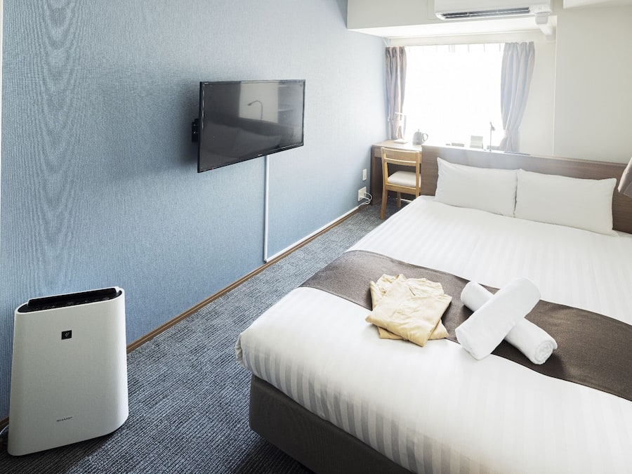 Quarto de hotel com cama, escrivaninha, cadeira, televisão e janela com cortinas.