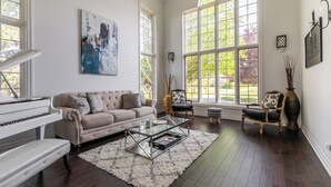 A spacious living room with a grand piano, a beige sofa, a glass coffee table, and large windows offering a view of the outdoors.