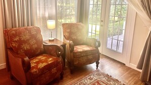 Une pièce avec deux fauteuils, une petite table avec une lampe et une grande fenêtre avec des rideaux.