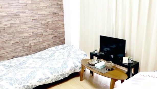 Ein Schlafzimmer mit einem Bett, einem Holzkopfstück, einem Fernseher auf einem Schrank, einem kleinen Tisch mit Tablett und einem Fenster mit Vorhängen.