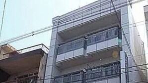 Ein mehrgeschossiges Gebäude mit Eingang im Erdgeschoss und einem Balkon im zweiten Stock. Ein Fahrrad steht davor geparkt.