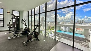 A gym with exercise equipment and a city view through large windows. - Stunning City Center Condo Full AC, Pool, Near All (Santo Domingo)