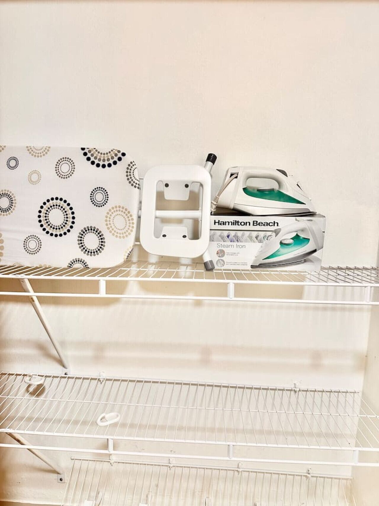 A neatly organized storage area with a patterned ironing board, a white ironing board, and a Hamilton Beach steam iron on a metal shelf.