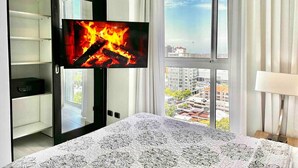 Ein Schlafzimmer mit einem Bett, einem Fernseher, der ein Feuer zeigt, einem Fenster mit Blick auf die Stadt und einem Kleiderschrank.