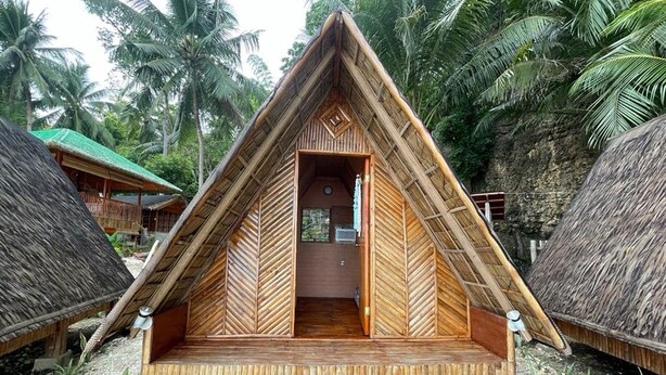 A thatched-roof, triangular-shaped hut with a wooden interior, surrounded by palm trees and other huts. - Hagdan Reef Dive Spot by HiveRooms (Oslob)