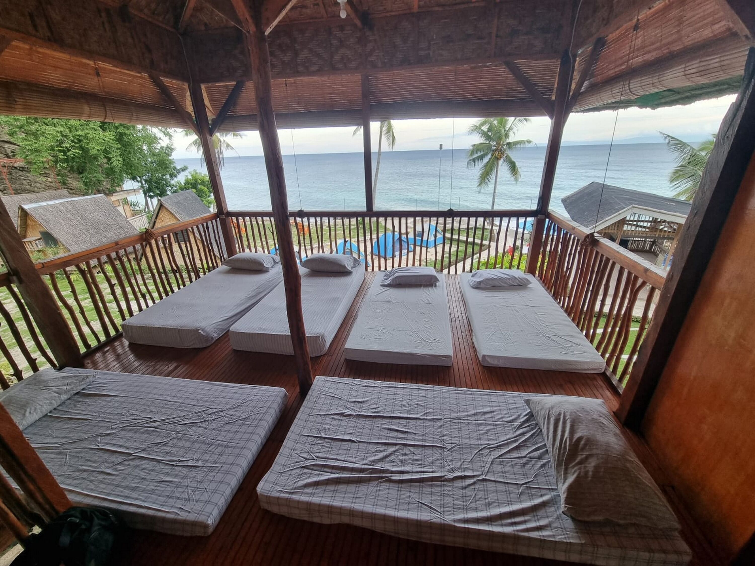 Eine Holzdielen-Terrasse mit vier Einzelbetten, jeweils mit Matratze und Kissen, mit Blick auf Strand und Meer.