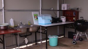 A compact kitchen with stainless steel sinks, a blue storage bin, a white refrigerator, and a barbecue grill. - Lucky Home Hotel and Apartment (Tinian)