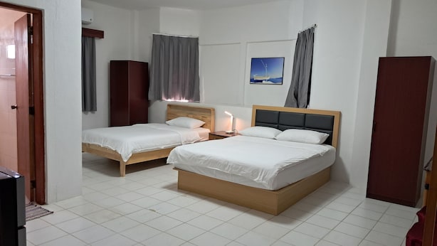 A hotel room with two single beds, a wooden headboard, a bedside table with a lamp, a wardrobe, and a painting on the wall. - Lucky Home Hotel and Apartment (Tinian)