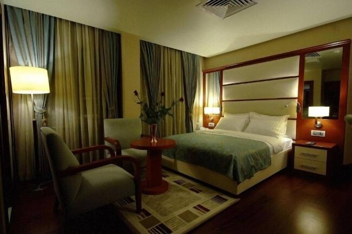 A hotel room with a bed, a chair, a table with a vase, and a nightstand with a lamp.