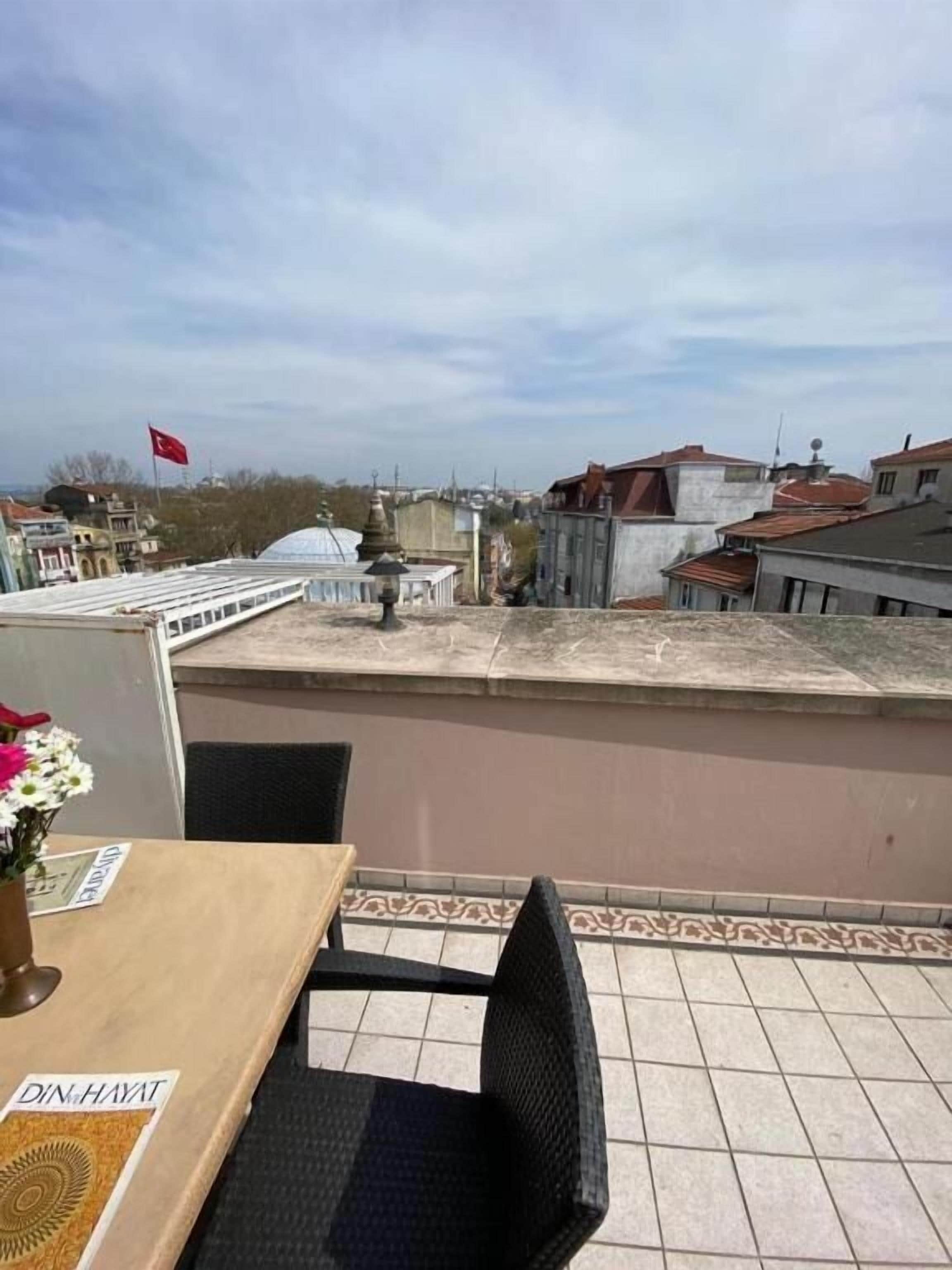 A rooftop terrace with a table and chairs, a flag, and buildings in the background.