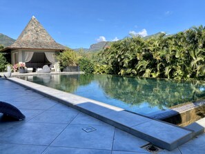 A swimming pool with a tiled deck, a covered seating area with cushions, and lush greenery in the background. - Villa Contemporaine - Côte Ouest (Rivière Noire)
