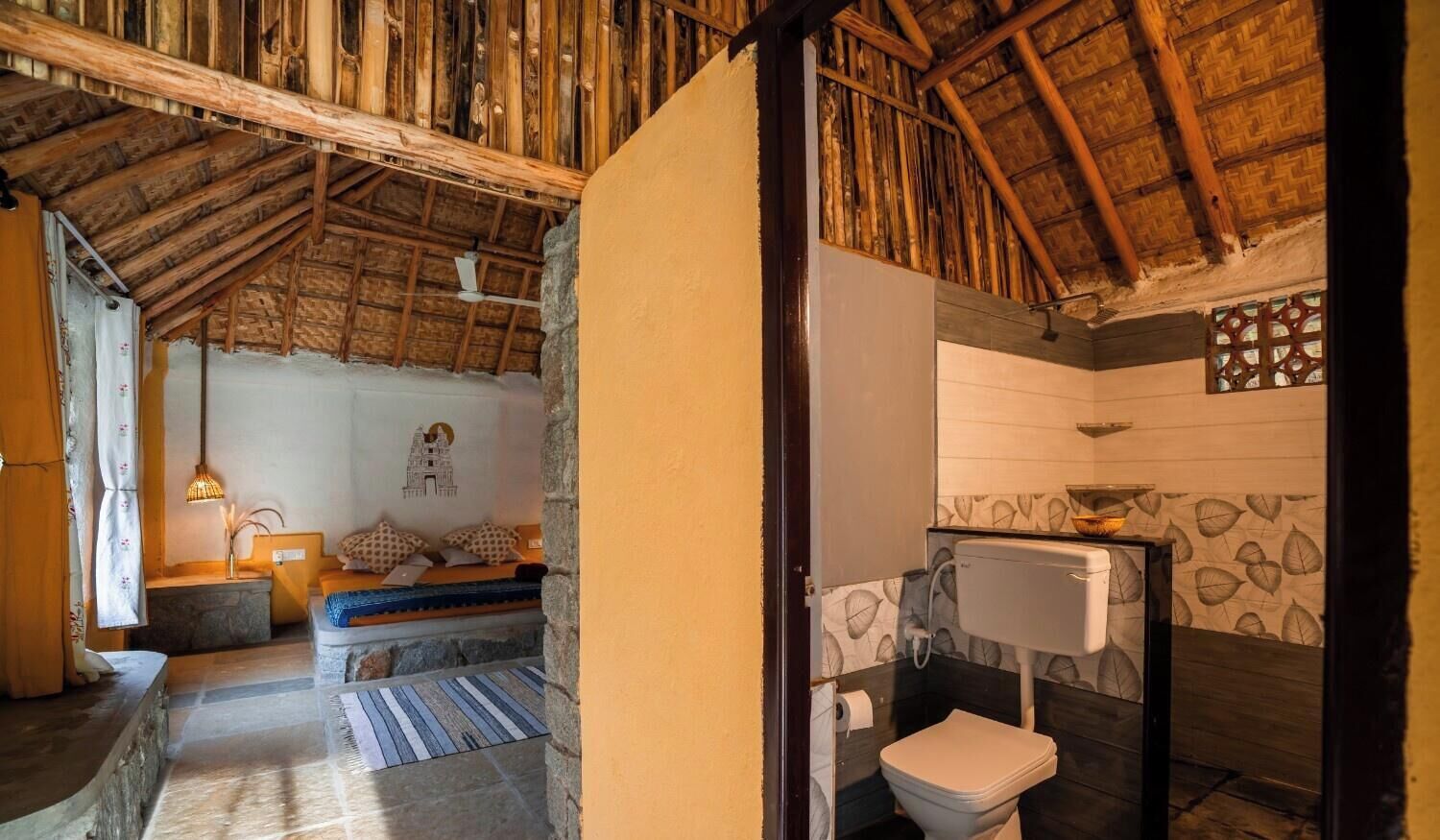 A rustic room with a thatched roof, a stone bench, a small table with a lamp, and a bathroom with a toilet and sink.