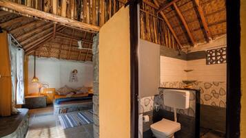 A rustic room with a thatched roof, a stone bench, a small table with a lamp, and a bathroom with a toilet and sink.