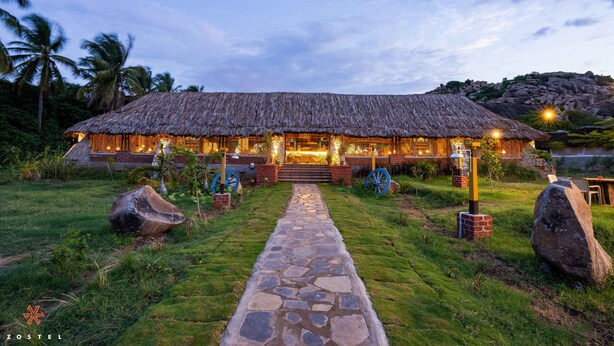 A thatched-roof building with a stone pathway leading to the entrance, surrounded by greenery and palm trees. - Zostel Hampi (Gangavathi) (Gangavathi)