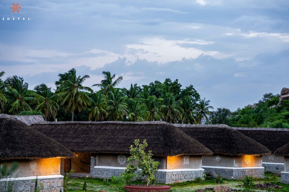 Thatched-roof huts with stone walls, arranged in a row, surrounded by lush greenery and palm trees.