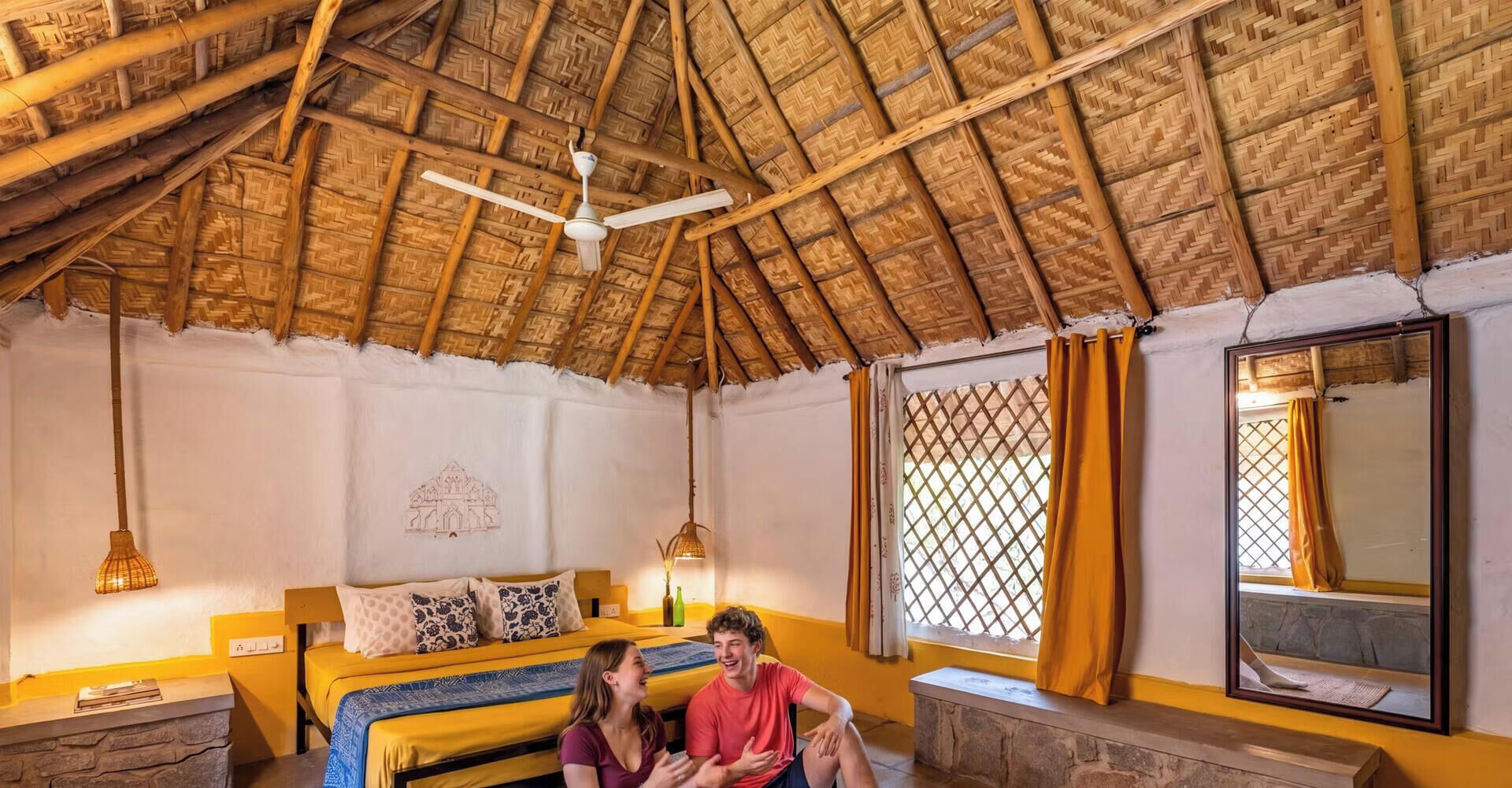 Una habitación con techo de paja, cama con almohadas estampadas, ventana con cortinas y un espejo montado en la pared.