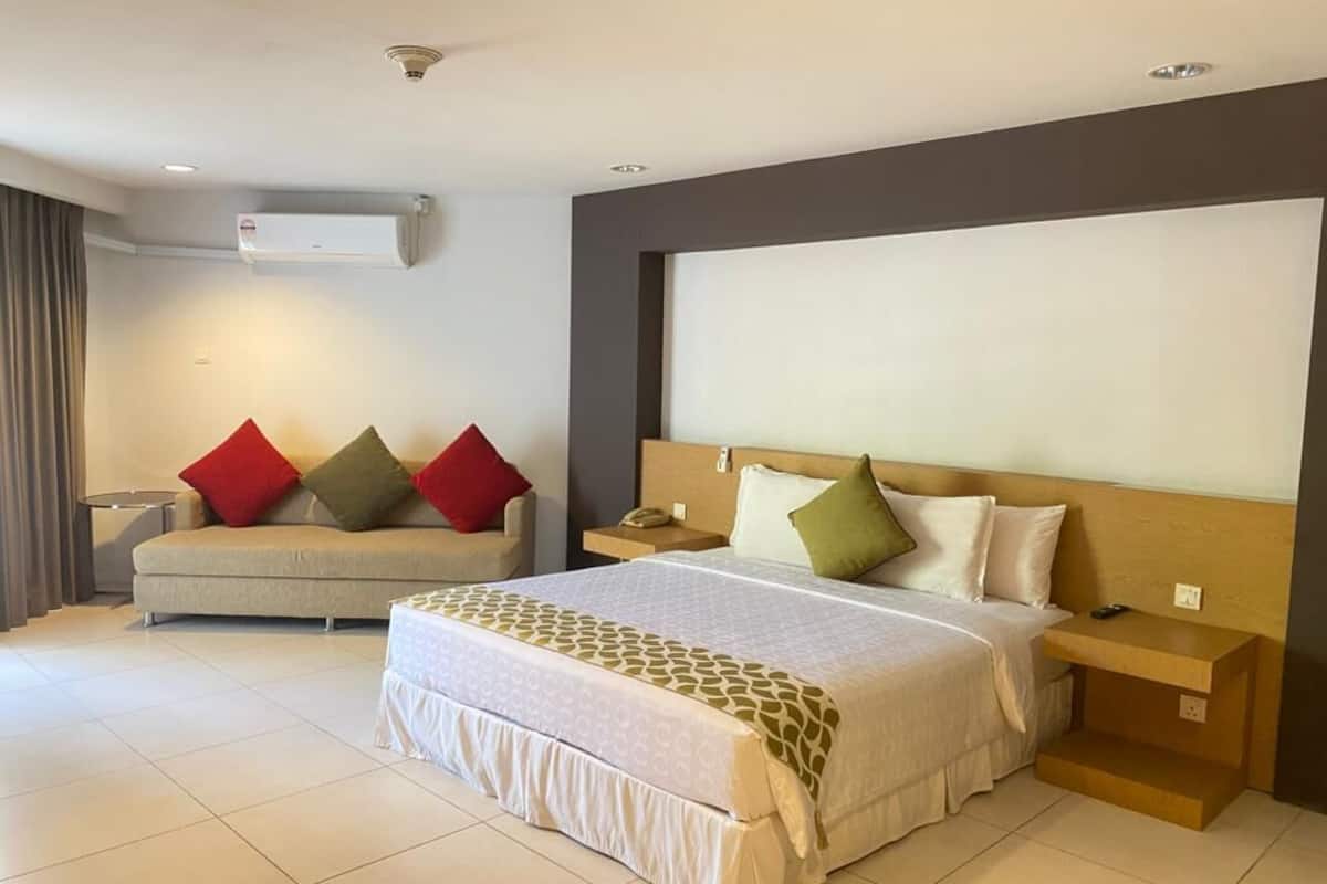 A hotel room with a large bed, a sofa, a nightstand, an air conditioning unit, and a wall-mounted TV.
