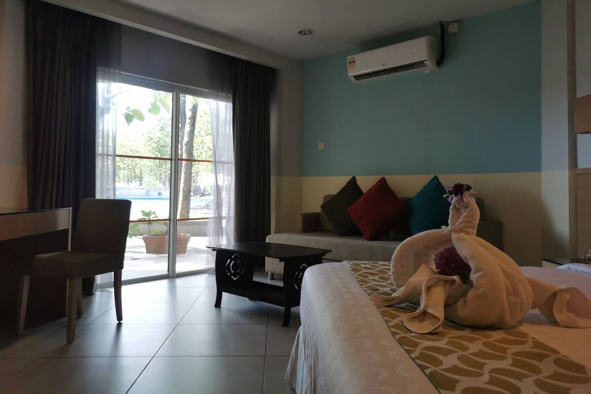 A hotel room with a bed, a chair, a table, an air conditioning unit, and a view of a pool through a glass door.