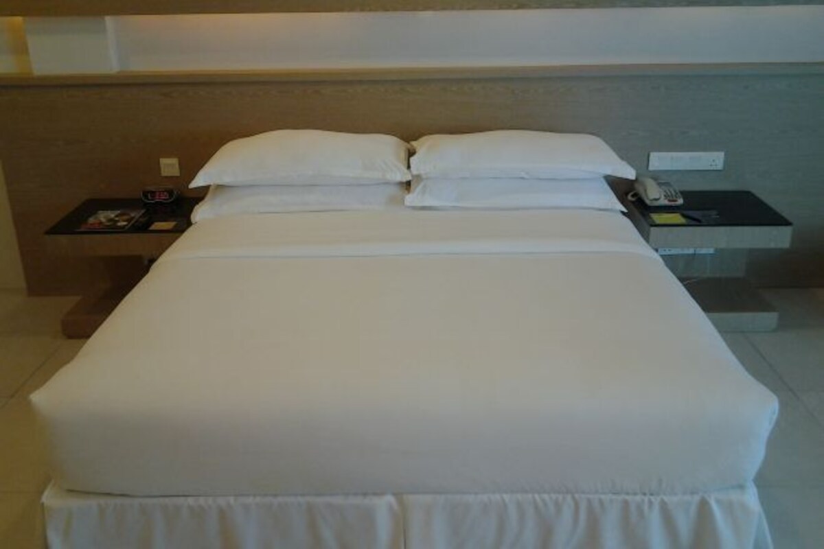 Habitación de hotel con una cama grande, dos almohadas, una mesita de noche con teléfono y una pequeña mesa con reloj y revista.