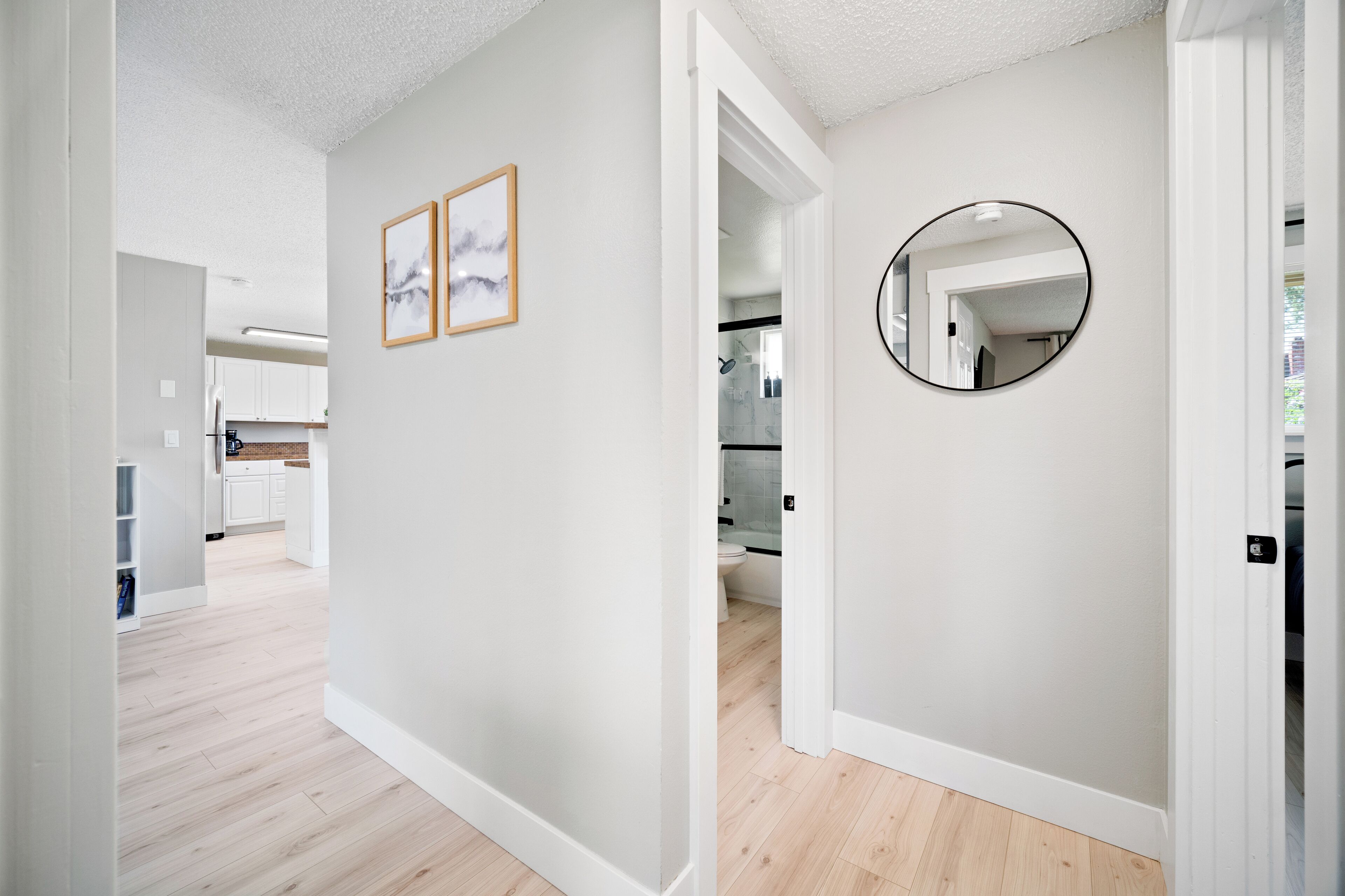 A hallway with light wooden flooring, white walls, and a round mirror on the right. There are framed pictures on the left wall.