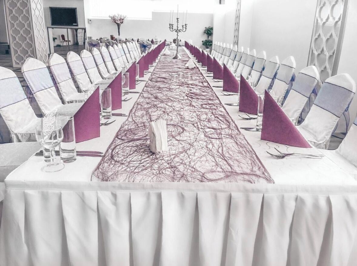 Una larga mesa dispuesta para un evento formal, con sillas blancas y servilletas moradas.