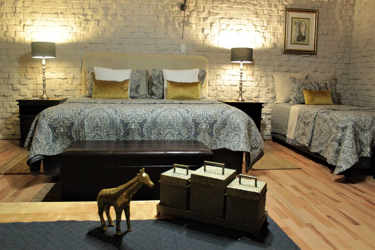 A bedroom with a brick wall, two beds with patterned bedding, bedside lamps, a wooden floor, and a small decorative animal.