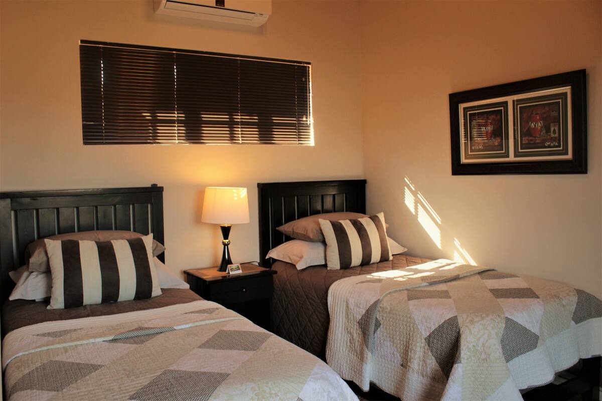 A hotel room with two beds, a nightstand with a lamp, a window with blinds, and two framed pictures on the wall.