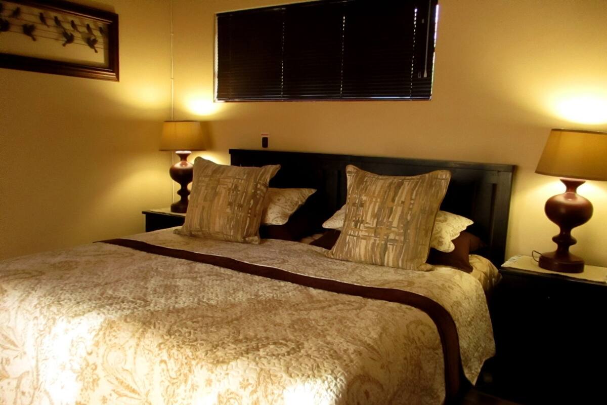 A neatly made bed with a patterned bedspread and pillows, flanked by two bedside tables with lamps, in a room with a window and a framed picture on the wall.