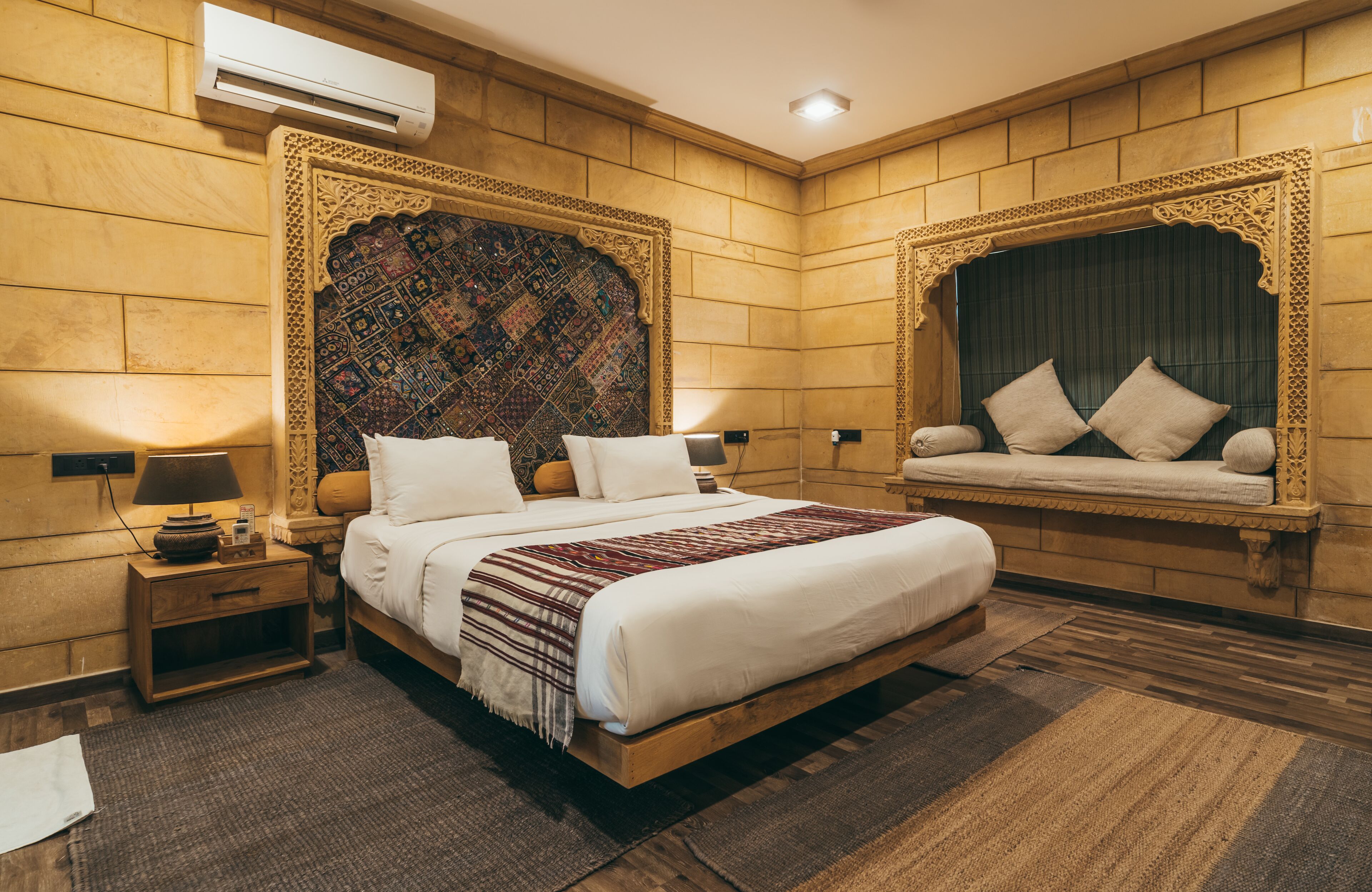 A bedroom with a large bed, a headboard with intricate patterns, a window seat with cushions, and a wall-mounted air conditioner.