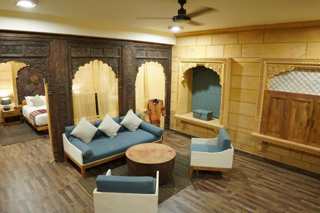 A room with a blue sofa, two white armchairs, a round wooden coffee table, and a bed with a white and red blanket.