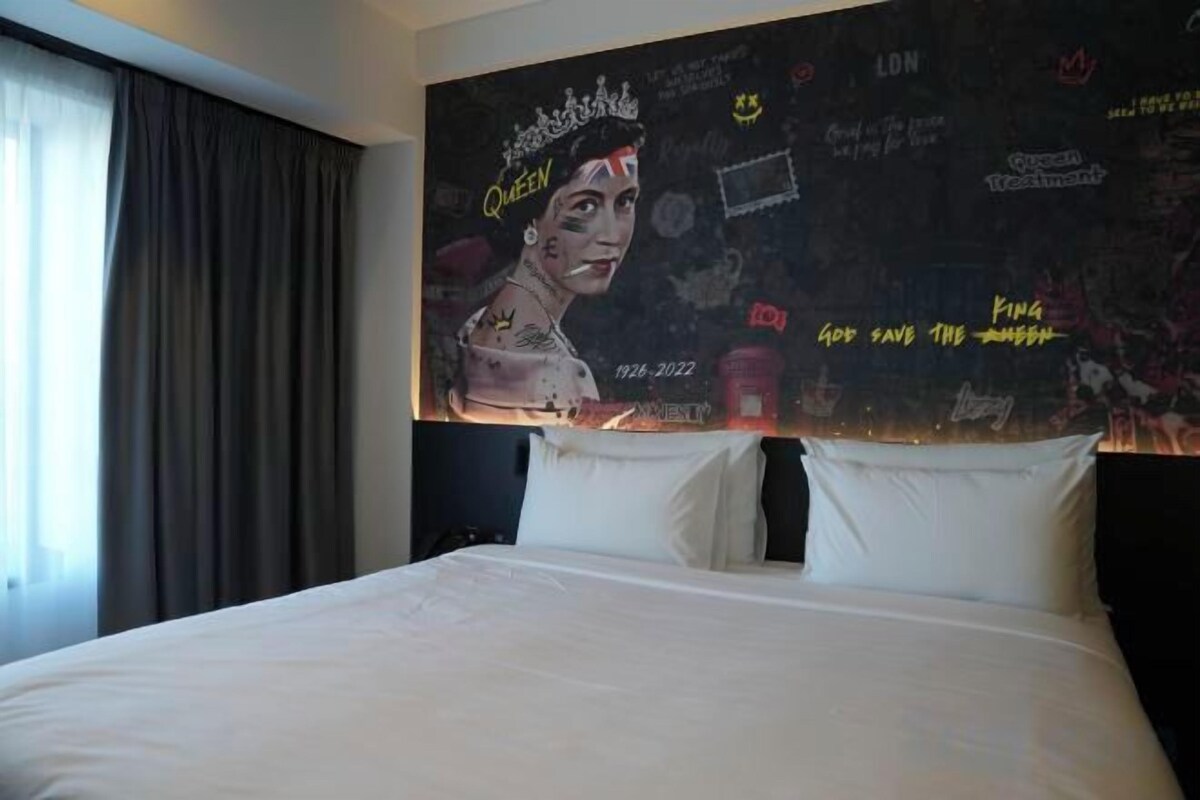 A hotel room with a large bed, a headboard with artwork, and a window with curtains.