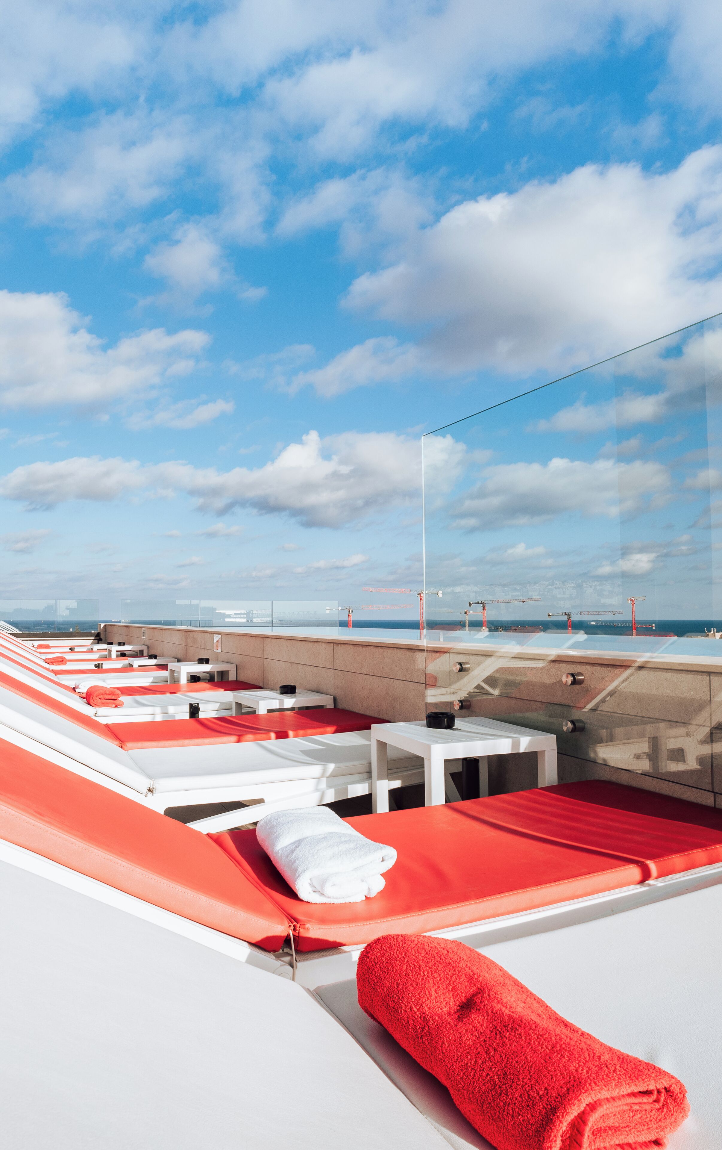 Eine Dachterrasse mit roten und weißen Lounges, einer Glasreling und Blick auf das Meer sowie Baukräne.