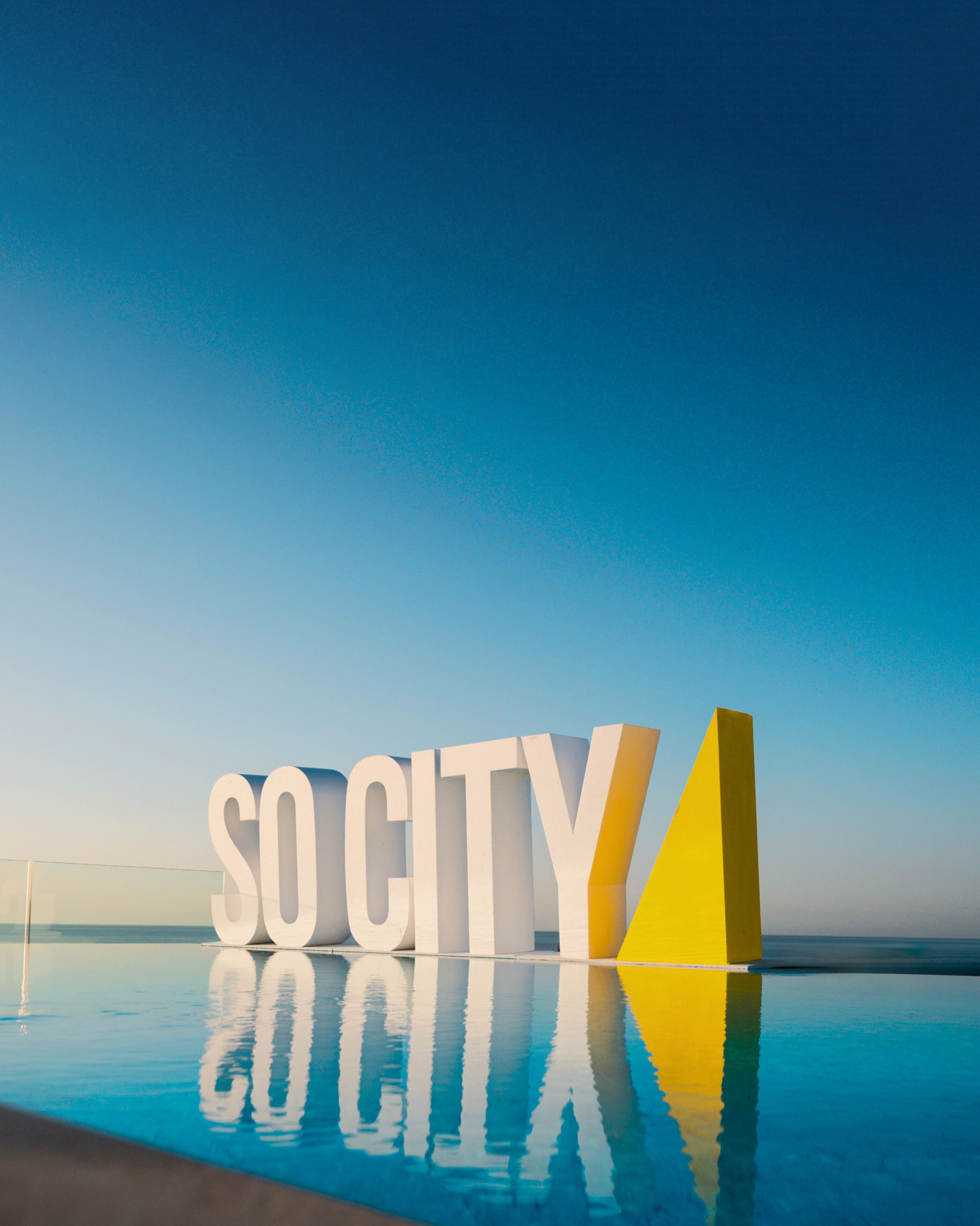 Grandes letras blancas que dicen "SO CITY" sobre la superficie reflectante de una piscina, con un cielo azul despejado al fondo.