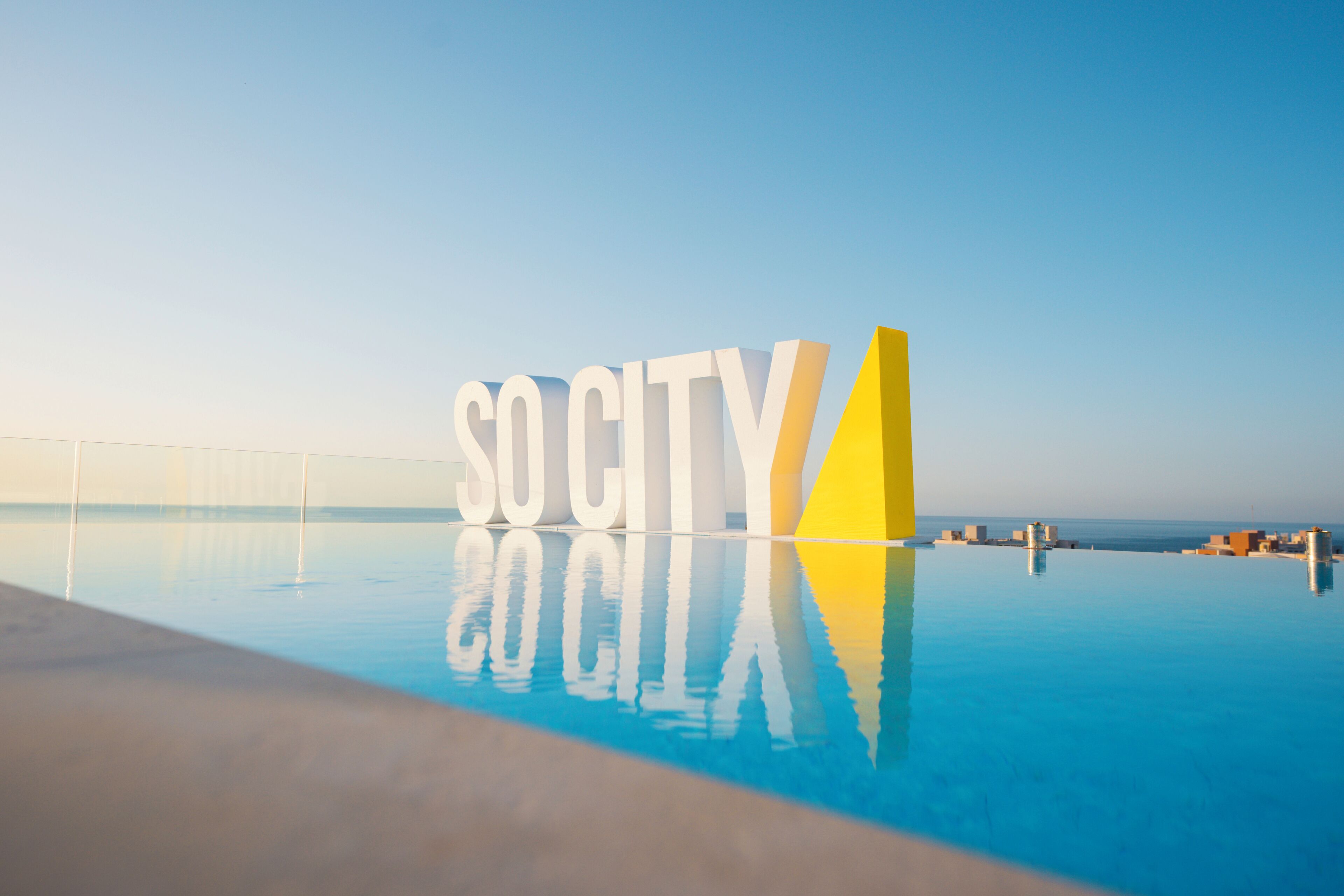 Une piscine dans laquelle le mot « SOCIÉTÉ » est inscrit en grandes lettres, entourée d’un ciel bleu clair et offrant une vue lointaine sur des bâtiments.